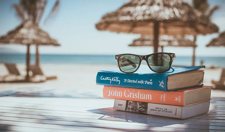 A pair of sunglasses is on top of books by famous authors on a beach table.