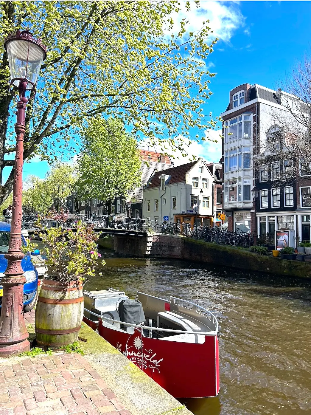 A scenic Amsterdam canal with a red boat docked near a lamppost, charming multi-story buildings, and a bicycle-lined bridge.